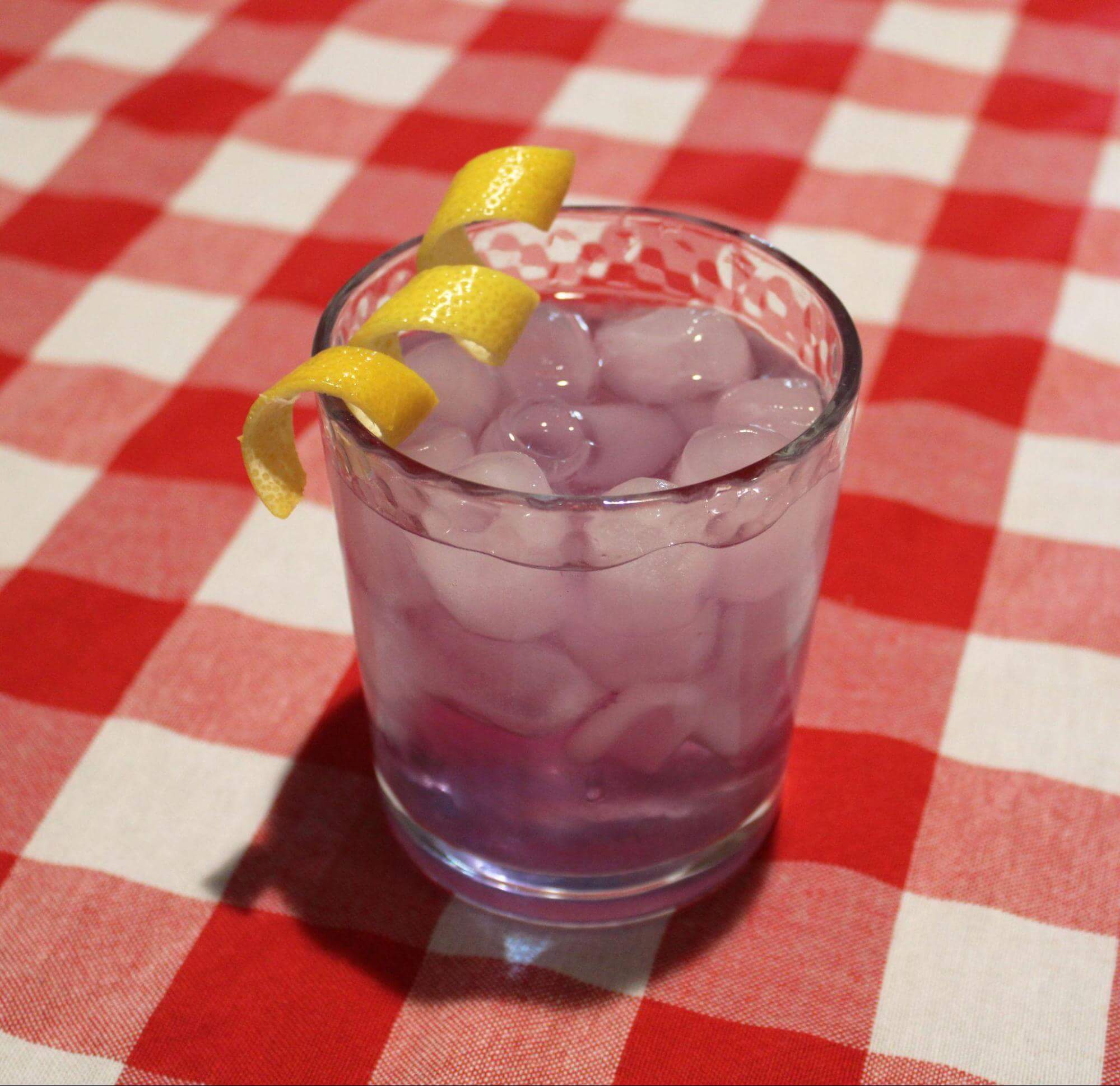 Glass of The Lavendar Menace sitting on red checkered picnic table cloth with lemon twist on the rim.