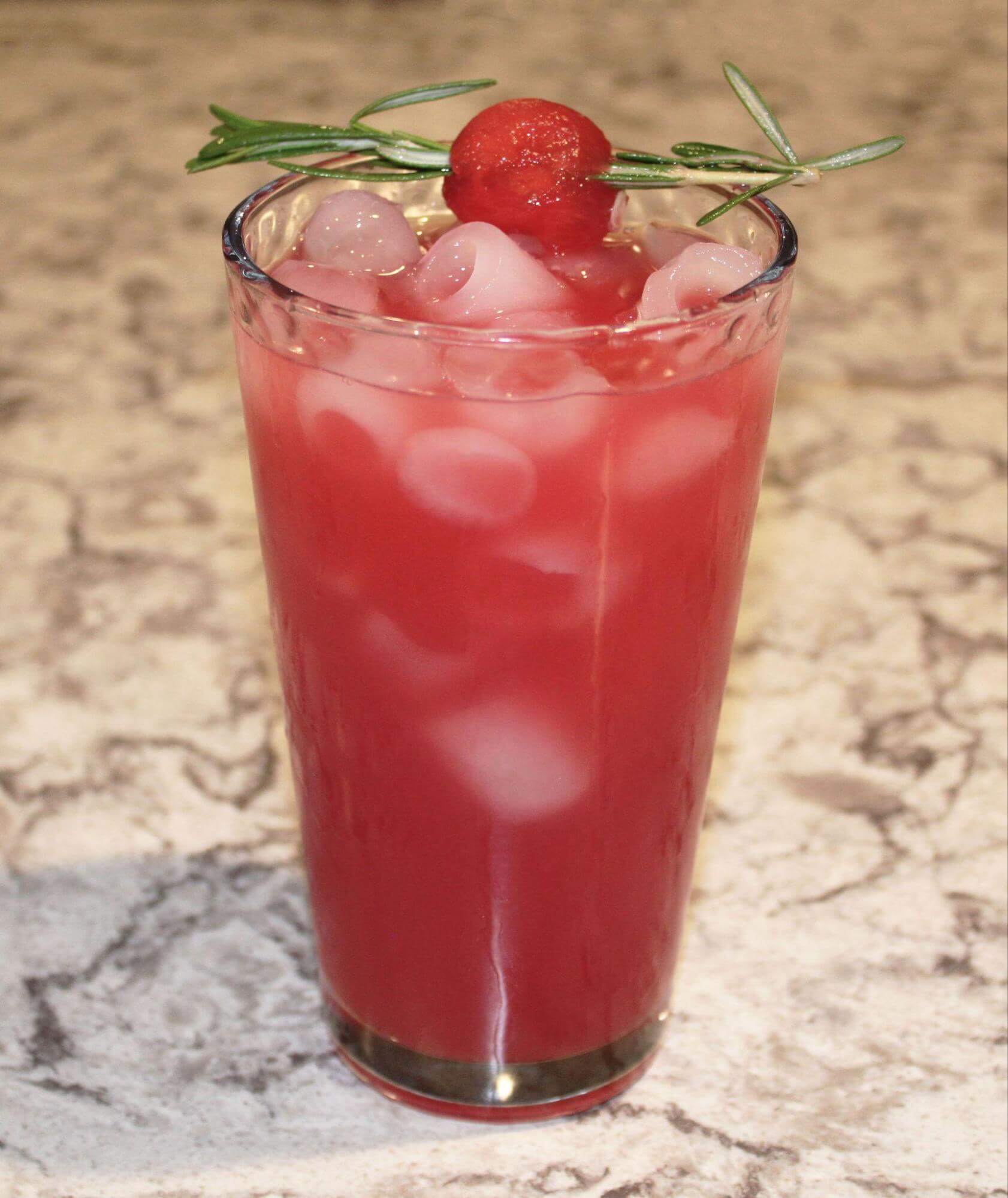 Tall glass of Watermelon Moon, a light red drink poured over ice, with a garnish of watermelon pierced with rosemary sitting across the top.